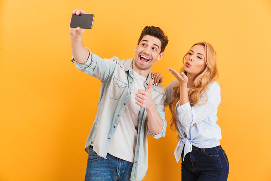 Portrait of two attractive people man and woman taking selfie photo on cell phone while gesturing at camera, isolated over yellow background