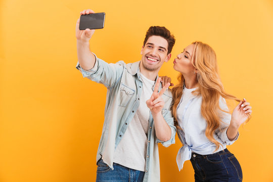 Portrait of lovely couple taking selfie photo on cell phone while woman kissing man on his cheek, isolated over yellow background