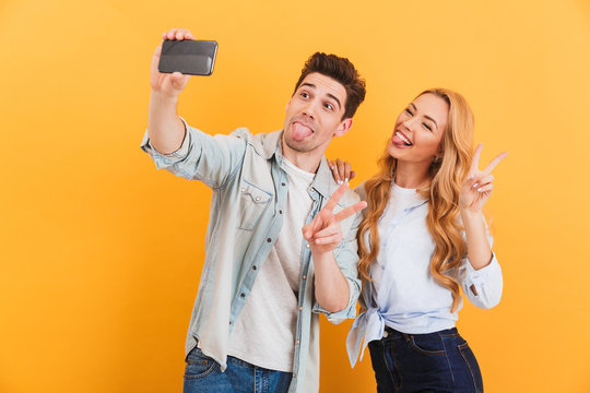 Portrait of two funny people man and woman taking selfie photo on smartphone while showing victory sign, isolated over yellow background