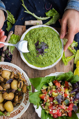 Woman hands holds avocado spinach green spread, dip Mix sauce with spoon. With basil leaves, ready for lunch or dinner with backed potato and vegetables salad. raw vegan vegetarian healthy food.