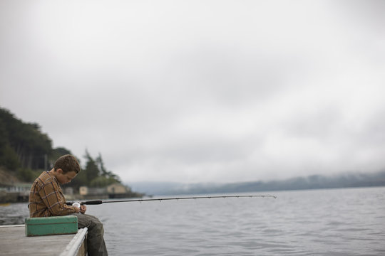 Unhappy boy fishing alone off the edge of a wooden jetty with a fishing rod.
