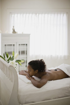 Young woman lying in bed on her stomach with healing stones on her back inside her bedroom.