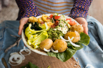 Woman hands holding buddha bowl with backed potato, seaweed, vegetables, spinach leaves, pesto dip sauce, salad, garlic. Raw vegan vegetarian healthy food on lunch or dinner