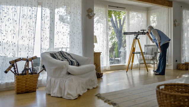 Mature woman looking through a telescope from her living room.