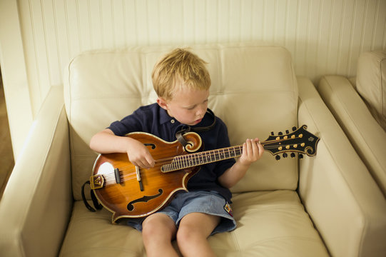 Young boy sitting in an armchair plays a small acoustic eight-string guitar.