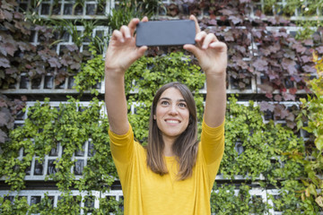 portrait outdoors of a beautiful young woman taking a picture with mobile phone. Wearing a yellow casual shirt and smiling. LIfestyle and fun. Green background