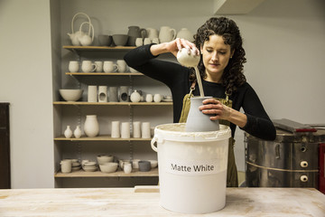 Woman with curly brown hair wearing apron standing in pottery workshop, pouring white glaze over unfired vase.