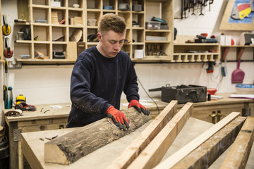 Man wearing work gloves standing at a workbench in a workshop, sanding bark of a piece of tree trunk.