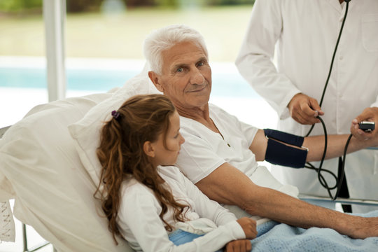 Man having his blood pressure taken in hospital, while receiving a visit from his granddaughter.