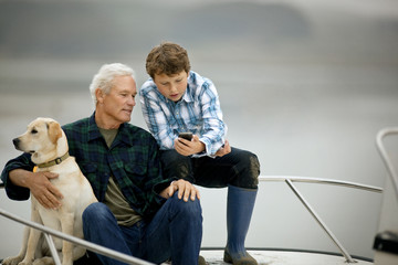 Man and his grandson sitting on the deck of a boat and looking at something on a cellphone.