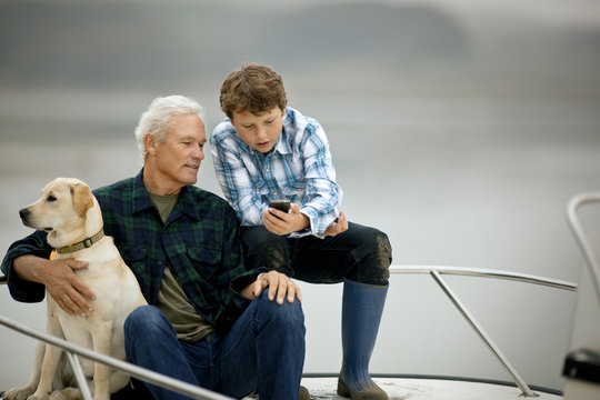 Man and his grandson sitting on the deck of a boat and looking at something on a cellphone.