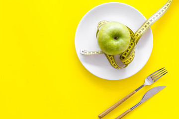 Proper nutrition with dietary fibre for weight loss. Apple on plate near measuring tape on yellow background top view copy space