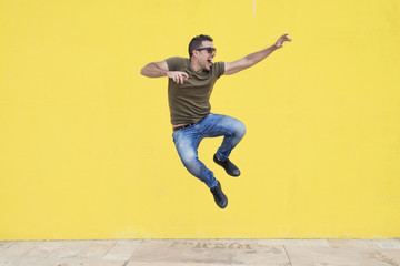 Young man with sunglasses jumping in front of a yellow wall.