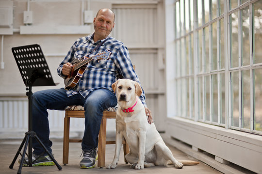 Portrait of a mid-adult man holding a guitar and petting his dog while sitting in a chair.