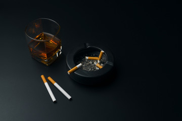 a glass of bourbon whiskey, cigarettes and black ceramic ashtray full of ashes with the cigarette butt, on the black table