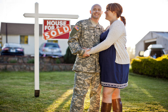 Smiling soldier standing with his pregnant wife next to a real estate sign in the front yard of their home.