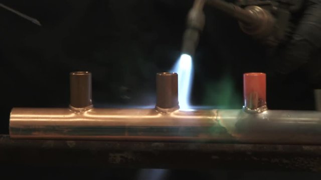 Person heats up copper tubing with blow torch, soldering metals together making custom product in warehouse.