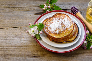 Sweet homemade pancakes with powdered sugar on a plate, decorated with flowers of an apple tree on a wooden background.