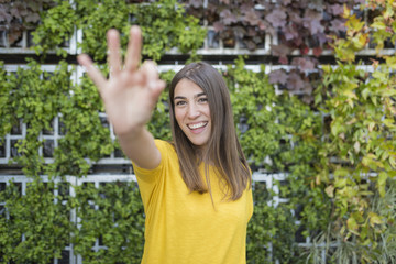 portrait outdoors of a beautiful young woman making the ok sign and smiling. Wearing a yellow casual shirt over green background. LIfestyle and fun.