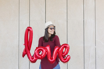 beautiful young woman having fun outdoors with a red balloon with a love word shape. Casual clothing. wearing hat and modern sunglasses. LIfestyle outdoors