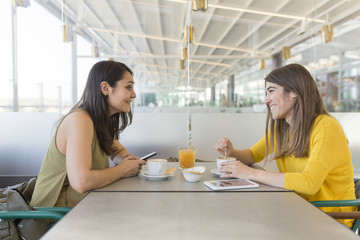 two beautiful women having breakfast in a restaurant. They are laughing and searching information on a mobile phone. Indoors lifestyle and friendship concept