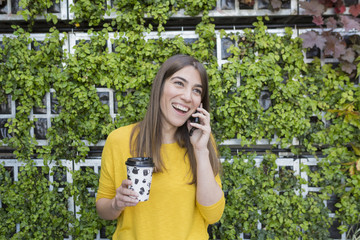 portrait outdoors of a beautiful young woman holding a cup of coffee, talking on her mobile phone and smiling. Wearing a yellow casual shirt over green background. LIfestyle and fun.