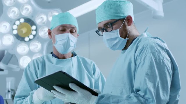 Two Professional Surgeons Use Digital Tablet Computer while Standing in the Modern Hospital Operating Room. Shot on RED EPIC-W 8K Helium Cinema Camera.