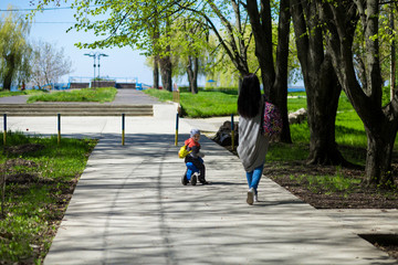 Young mother walks with small son outdoors with children's motorbike. Active childhood