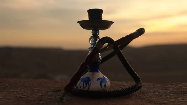 Hookah, traditional arabic waterpipe, direct sunset light. Outdoor slider shot. Silhouettes of hookah on sunset mountain background. Hookah hot coals on shisha bowl at the desert. Eastern shisha loung