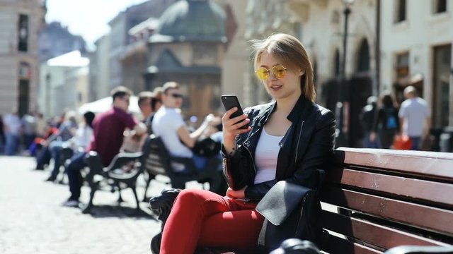 happy charming young woman with blonde hair looking at smartphone screen sitting on bench outside laughing surprised positive emotional face checking message notification day sunny light sunshine
