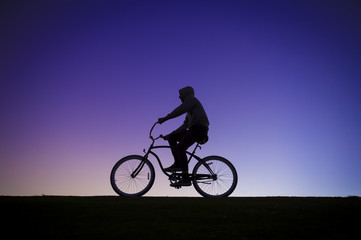 Fototapeta na wymiar Silhouette of a man in a hoodie riding a cruiser bike in front of a purple sunset sky