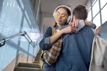 young stylish female tourist in sunglasses with headphones and smartphone hugging boyfriend at subway