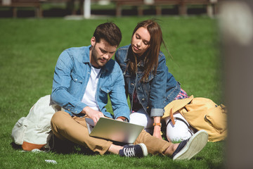 young tourists with backpacks and laptop sitting on grass