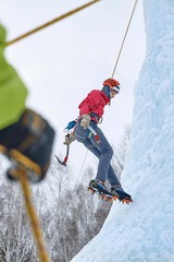 Alpinist woman with  ice tools axe in orange helmet climbing a large wall of ice. Outdoor Sports...