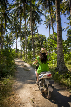 young happy tourist woman with hat riding scooter motorbike in tropical paradise jungle  with blue sky and palm trees exploring trip destination