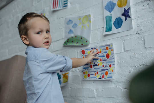 little kid looking at camera while hanging childish pictures on wall at home
