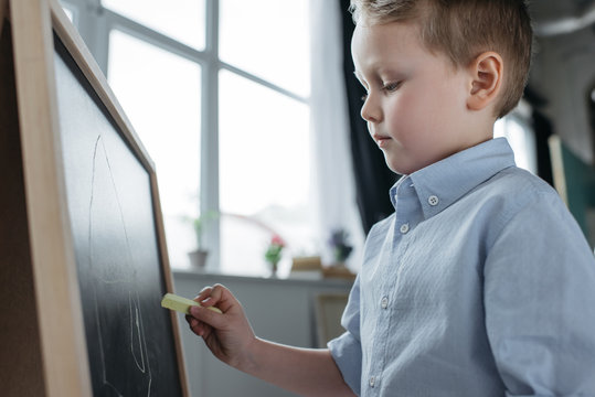 side view of little boy with piece of chalk drawing picture on blackboard at home