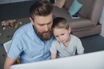 father and little son looking at computer screen while using computer at home