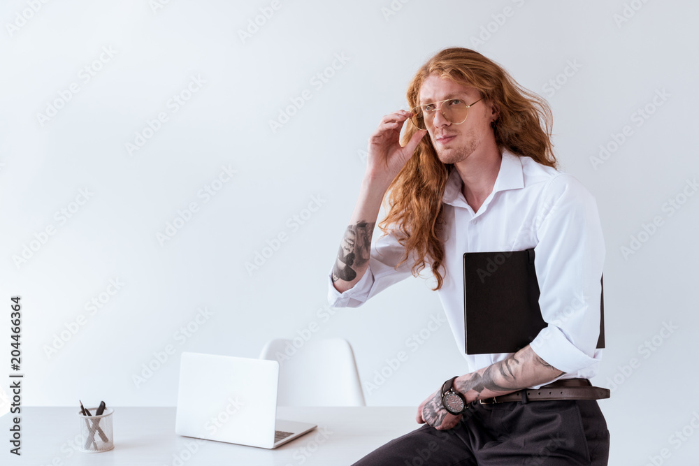 Wall mural stylish tattooed businessman with curly hair sitting on table and touching glasses in office