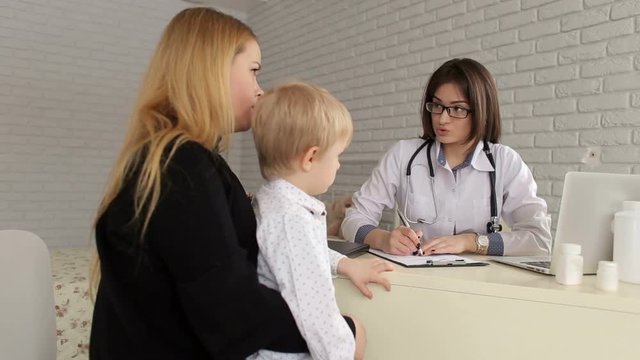 Woman doctor talking to the mother of a young boy and makes notes in a notebook, they sit in the office at the table. Pediatrics. Family doctor.