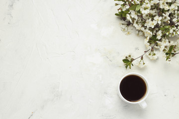 cup with black coffee, branches with white flowers on a light stone background. Flat lay, top view