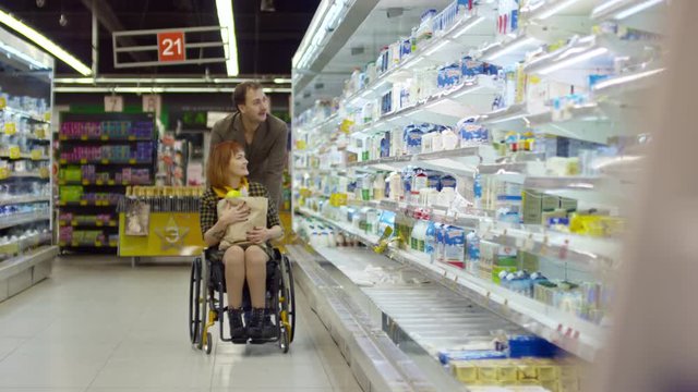 Loving husband pushing young paraplegic woman in wheelchair while shopping in dairy department in supermarket