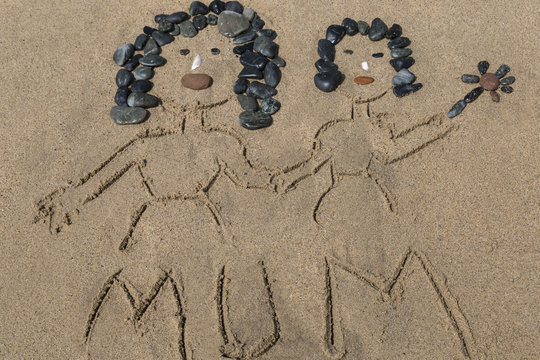 drawing a picture with stones of mother's day on sand on the beach. kid holds the flower and her mum's hand.