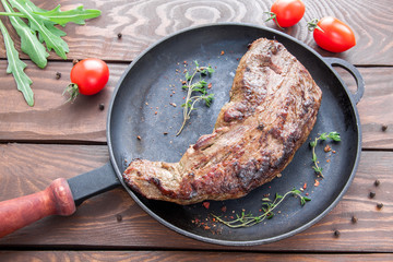 roast beef fillet with a delicious crust on a black iron frying pan with seasonings and herbs and a twig of twig on a wooden table with red ripe tomatoes and green arugula