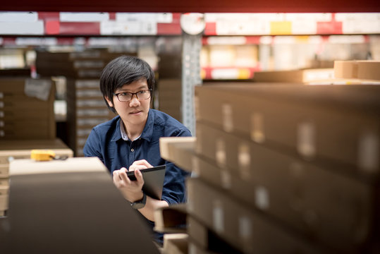Young Asian man doing stocktaking of product in cardboard box on shelves in warehouse by using digital tablet. physical inventory count concept