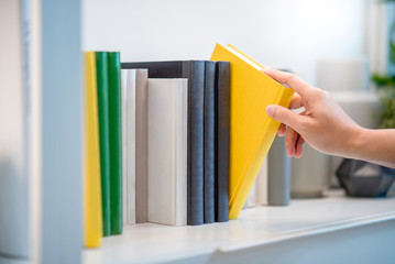 male hand choosing and picking white book on white bookshelf in public library, education research...