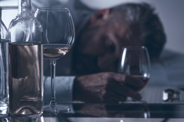 Close-up of a drunken desperate middle-aged man leaning on the table behind bottles and glasses with alcohol
