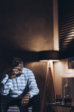Silhouette of a stressed middle-aged man with problems sitting alone with his head down next to a bottle and a glass of alcohol in a dark room with window blinds and a dimmed light of a standing lamp