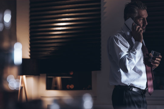 Tired and sad middle-aged businessman talking on the phone and holding a glass with alcohol while standing in a room with dimmed light and window blinds