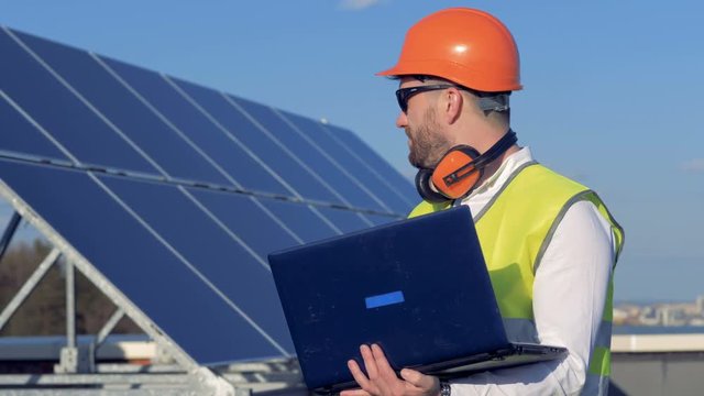 Worker uses his laptop, while examining the solar panels on the roof. 4K.